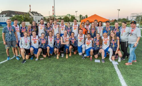 Eine große gemischte Sportmannschaft in passenden Uniformen posiert auf einem Sportplatz, lächelnd und mit Medaillen. Einige Leute knien vor ihnen, während andere hinter ihnen stehen. Im Hintergrund sind Gebäude und ein orangefarbenes Zelt zu sehen.