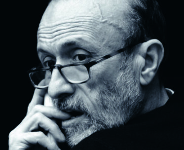 Ein Mann mit Brille und Bart blickt über seine Schulter und stützt seine Hand auf den Mund. Das Bild ist in Schwarz-Weiß gehalten, was die Linien und die Textur seines Gesichts betont.