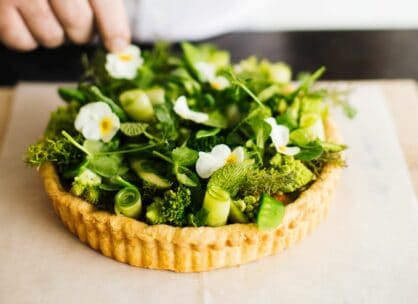 Eine Hand fügt essbare Blumen und frisches grünes Gemüse zu einer offenen Tarte mit goldener Kruste hinzu, die auf Pergamentpapier angerichtet ist.