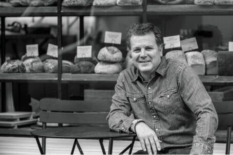 Ein Mann in einem Jeanshemd sitzt an einem Tisch im Freien vor dem Schaufenster einer Bäckerei, in der verschiedene Brotsorten ausgestellt sind. Er lächelt und schaut in die Kamera. Das Bild ist in schwarz-weiß gehalten.