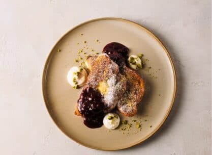 Ein beiger Teller mit zwei Scheiben French Toast, belegt mit Puderzucker, dunkler Beerensauce, Schlagsahne und gehackten Pistazien auf hellgrauem Hintergrund.