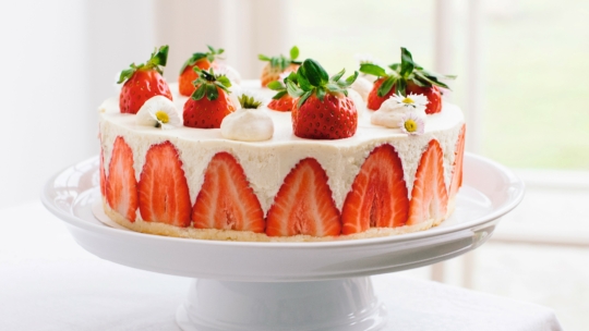 Ein runder Erdbeerkuchen auf einem weißen Tortenständer, verziert mit halbierten Erdbeeren an den Seiten und ganzen Erdbeeren und kleinen weißen Blumen obenauf. Die Torte wird in einem hellen Raum mit natürlichem Licht aufgestellt.