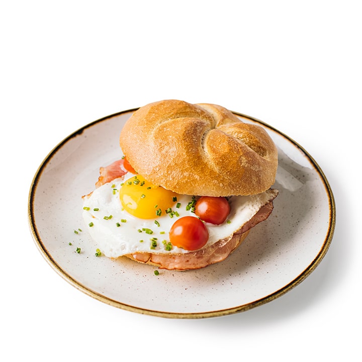 Ein Sandwich auf einem runden Brötchen mit Schinken, einem Spiegelei, halbierten Kirschtomaten und gehacktem Schnittlauch, serviert auf einem weißen Teller mit braunem Rand auf weißem Hintergrund.