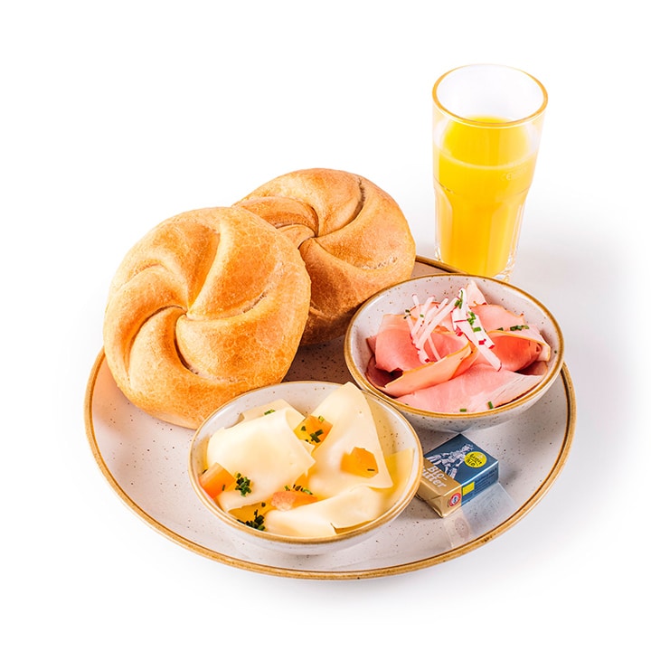 Ein Frühstücksteller mit zwei Brötchen, einer Schale mit Schnittkäse, einer Schale mit Schinken und Radieschen, einer kleinen Butterpackung und einem Glas Orangensaft, alles auf einem weißen Teller angerichtet.