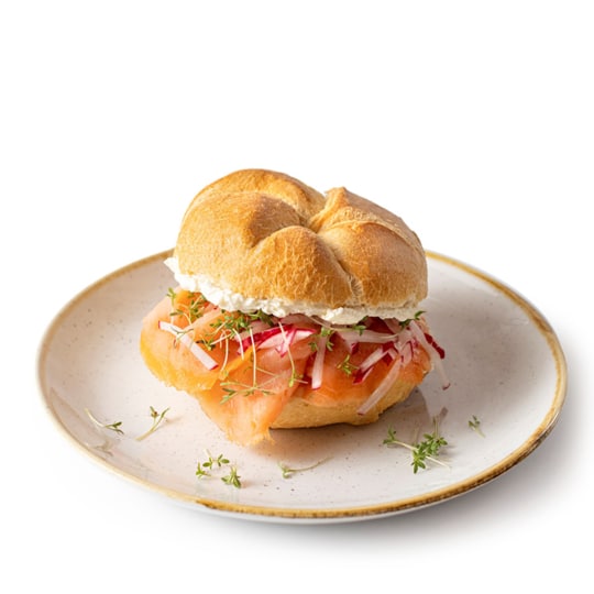 Ein rundes Sandwichbrötchen, gefüllt mit Räucherlachs, Frischkäse, Radieschenscheiben und frischen Sprossen, serviert auf einem beigen Teller vor weißem Hintergrund.