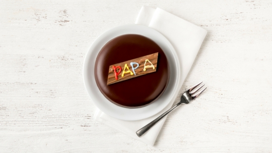 Das Vatertagstörtchen, ein runder schokoladenglasierter Kuchen auf einem weißen Teller, gekrönt von einer holzähnlichen Plakette mit der Aufschrift PAPA in bunter Glasur. Eine Gabel und eine gefaltete Serviette liegen neben dem Teller auf einer weißen Holzfläche.