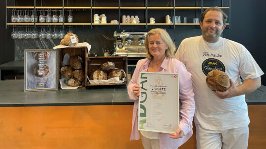 Eine Frau mit einer Urkunde und ein Mann mit einem Brot in der Hand stehen vor einer Theke mit mehreren runden Broten. Im Hintergrund sind Regale mit Gläsern und eine Kaffeemaschine zu sehen.