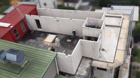 Luftaufnahme eines im Bau befindlichen Gebäudes mit teilweise fertiggestellten Betonwänden. Zwei Arbeiter sind im Inneren des Gebäudes zu sehen, und Baumaterialien liegen auf dem unfertigen Boden verstreut. Angrenzende Dächer sind sichtbar.