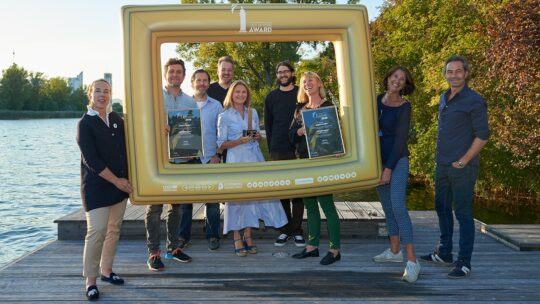 Eine Gruppe von neun Personen steht auf einem Holzsteg an einem See und hält einen großen aufblasbaren goldenen Bilderrahmen. Zwei Personen zeigen gerahmte Auszeichnungen. Im Hintergrund sind Bäume und Wasser an einem sonnigen Tag zu sehen.