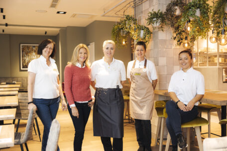 Fünf Frauen, einige in Freizeitkleidung, andere in Restaurantuniformen, stehen und sitzen lächelnd in einem modernen, gut beleuchteten Restaurant mit Holztischen und Hängepflanzen.