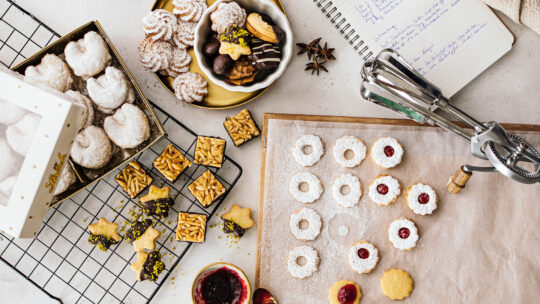 Ein Sortiment von Keksen und Gebäck auf einem Tisch, darunter mit Marmelade gefüllte Kekse, mit Puderzucker bestäubte Kekse und Kekse mit Nüssen. Ein handgeschriebenes Rezeptbuch und Backwerkzeuge sind ebenfalls zu sehen.