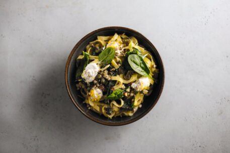 Eine Schüssel mit Nudeln, die mit frischem Blattspinat, Käse, Kapern und Kräutern auf einer hellgrauen, strukturierten Oberfläche angerichtet sind.