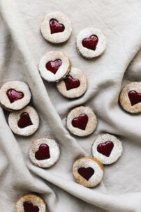 Herzförmige Linzer Kekse mit Puderzucker und sichtbaren roten Marmeladenkernen sind auf einem beigen Tuch verstreut und wirken so gemütlich und einladend.