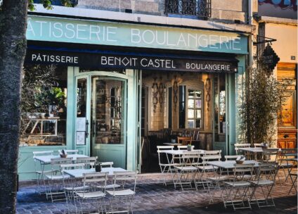 Sitzgelegenheiten im Freien vor einer französischen Bäckerei und Konditorei namens Benoit Castel mit weißen Tischen und Stühlen auf einem Kopfsteinpflaster und einer blaugrünen Schaufensterfront mit großen Fenstern.