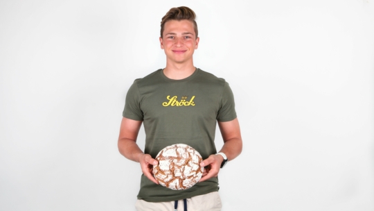 Eine junge Person mit kurzen Haaren lächelt, während sie einen runden Laib rustikalen Brotes in der Hand hält. Sie trägt ein grünes T-Shirt mit gelber Aufschrift und helle Shorts, die vor einem schlichten weißen Hintergrund stehen.