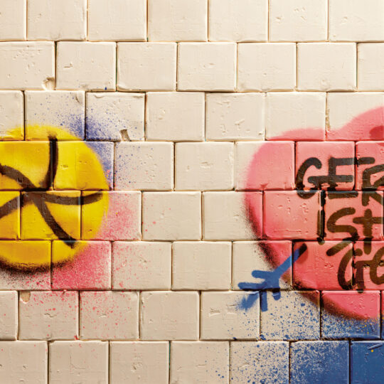 Das Graffiti auf einer gekachelten Wand zeigt einen gelben Kreis mit einem schwarzen Symbol und einem rosa Herz mit einem Pfeil, blauen Akzenten und den deutschen Worten "GERM IST GEIL".
