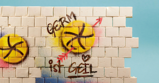 Eine Wand aus weißen Würfeln ist mit gelben Kreisen, schwarzen Linien und Wörtern in deutscher Sprache, darunter "GERM" und "ist GEIL", bemalt. Ein roter Pfeil weist auf den Text hin. Der Hintergrund ist hellblau.
