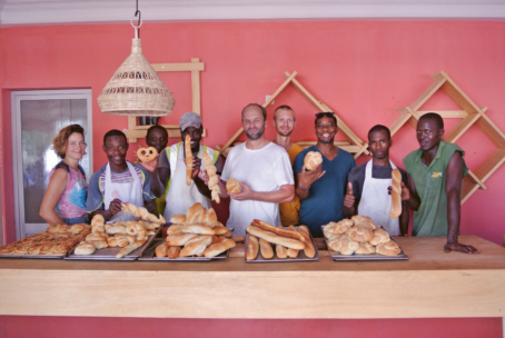 Eine Gruppe von Menschen steht hinter einer Theke, die mit verschiedenen Broten und Brötchen gefüllt ist, und lächelt in die Kamera in einer Bäckerei mit einer rosa Wand und dekorativen Holzregalen im Hintergrund.