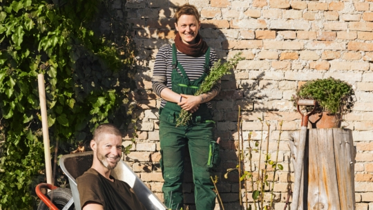 Eine Frau in grünem Overall hält Kräuter in der Hand und lächelt neben einer mit Efeu bewachsenen Backsteinmauer, während ein Mann in der Nähe in einer Schubkarre sitzt und ebenfalls lächelt. Um sie herum sind Gartengeräte und Pflanzen zu sehen.
