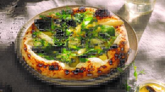 Eine Holzofenpizza mit frischem Blattgemüse, Kräutern und dünn geschnittenem Gemüse liegt auf einem Keramikteller, daneben ein Glas Wasser auf einem grauen Tischtuch.