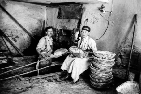 Zwei Männer sitzen in einem Haus und flechten Körbe; der eine flechtet, während der andere einen fertigen Korb hält. Neben ihnen liegen Stapel fertiger Körbe, und an der Wand hängen Werkzeuge in einem rustikalen, altmodischen Arbeitsraum.