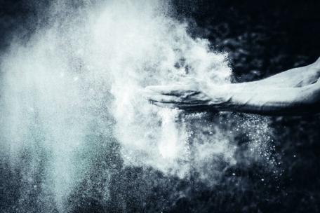 Eine Nahaufnahme von klatschenden Händen, die eine Wolke aus weißem Pulver freisetzen, vor einem dunklen, unscharfen Hintergrund. Das Pulver scheint entweder Kreide oder Mehl zu sein und erzeugt einen dramatischen, dynamischen Effekt.