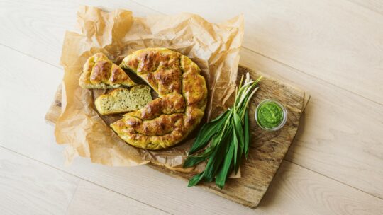 Ein runder Laib Kräuter-Focaccia auf Pergamentpapier, in zwei Scheiben geschnitten, auf einem Holzbrett neben frischen grünen Kräutern und einem kleinen Glas mit grünem Pesto.