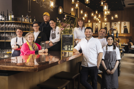 Eine Gruppe von Restaurantmitarbeitern, Köchen und eine Frau mit einem bunten Schal posieren gemeinsam lächelnd hinter einem polierten Bartresen mit Blumen, Auszeichnungen und Weingläsern in einem warm beleuchteten, modernen Restaurant.
