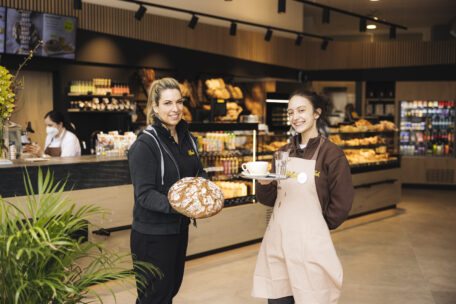 Zwei Frauen stehen in einer Bäckerei; die eine hält einen runden Laib Brot, die andere trägt ein Tablett mit einer Kaffeetasse und einem Glas Wasser. Im Hintergrund sind Regale mit Brot, Gebäck und Lebensmitteln zu sehen.