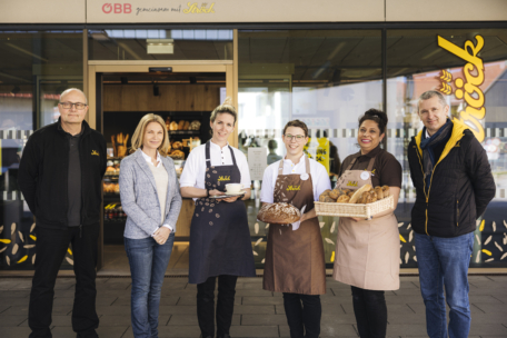 Sechs Personen stehen lächelnd vor einer Bäckerei; drei von ihnen tragen Schürzen und halten Backwaren in der Hand, während die anderen drei Freizeit- oder Geschäftskleidung tragen. Das Schaufenster und das Logo der Bäckerei sind im Hintergrund zu sehen.