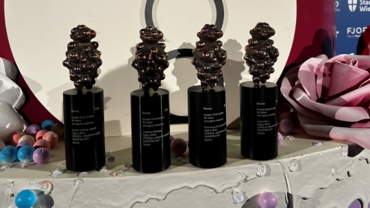 Vier Bronzetrophäen mit abstrakten, gruppierten Motiven sind auf einer dekorierten Fläche mit bunten Kugeln und künstlerischen Formen im Hintergrund ausgestellt. Jede Trophäe hat einen eingravierten Text auf einem schwarzen zylindrischen Sockel.