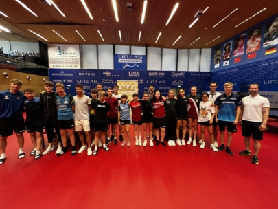 Eine Gruppe junger Sportler und zwei erwachsene Trainer stehen zusammen auf einem Tischtennisplatz und lächeln für ein Gruppenfoto. Im Hintergrund sind Sponsorenbanner und eine Wand mit Fotos und Fahnen zu sehen.