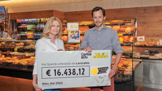 Zwei Personen stehen in einer Bäckerei und halten einen großen Spendenscheck über 16.438,12 € zur Unterstützung von Lerncafés in der Hand. Im Hintergrund sind Regale mit Brot und Gebäck zu sehen. Der Scheck ist datiert auf Mai 2022, Wien.