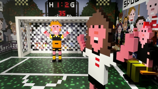 Verpixelte Fußballszene mit zwei blockigen Figuren: eine in Weiß feiert, eine in Gelb steht in der Nähe eines Tores. Die Anzeigetafel darüber zeigt 1:2 und im Hintergrund ist eine Menschenmenge abgebildet, alles im Retro-Videospiel-Stil.