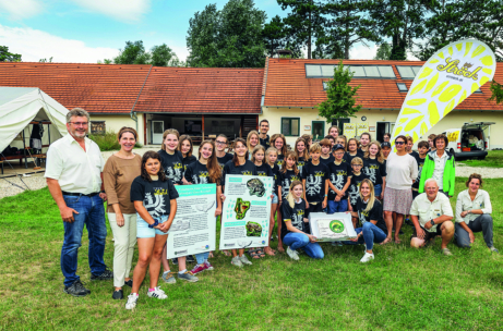 Eine Gruppe von Menschen, meist Jugendliche, posiert draußen vor einem Gebäude. Einige halten Informationsplakate in der Hand und tragen passende T-Shirts. Erwachsene stehen auf beiden Seiten, und rechts ist eine gelbe Flagge zu sehen. Die Szene ist hell und fröhlich.
