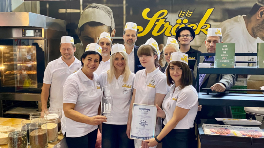 Eine Gruppe lächelnder Menschen in weißen Hemden und Hüten posiert zusammen in einem Innenraum bei einer Bäckereiveranstaltung und hält eine gläserne Trophäe und eine Urkunde in der Hand, während um sie herum Backgeräte und Zutaten zu sehen sind.