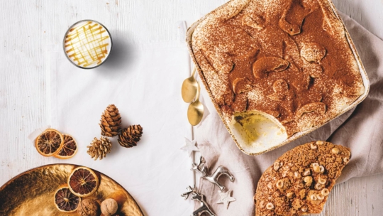 Eine quadratische Tiramisu-Schale mit einem fehlenden Stück liegt auf einer Serviette neben goldenen Löffeln, getrockneten Orangenscheiben, Tannenzapfen und einem goldenen Teller und sorgt für eine festliche und gemütliche Tischdekoration.
