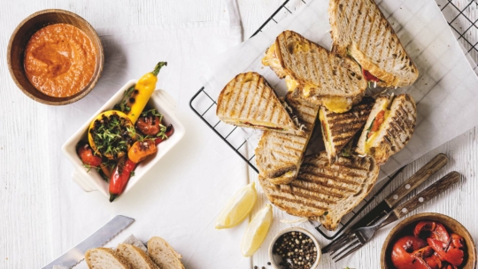 Gegrillte Sandwiches auf einem Kühlgestell mit Zitronenspalten, eine Schale Tomatensuppe, ein Salat mit Kirschtomaten und Paprika, Pfefferkörner, Brotscheiben und Besteck auf einem weißen Tisch.