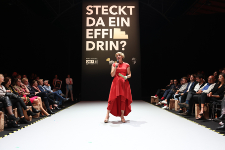 Eine Frau in einem roten Kleid spricht auf einer Laufstegbühne und wendet sich an ein Publikum, das auf beiden Seiten sitzt. Hinter ihr steht ein großes Schild mit der Aufschrift "STECKT DA EIN EFFIE DRIN?" in fetten Buchstaben.
