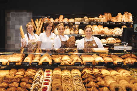 Vier Bäckereiangestellte stehen hinter einer mit Gebäck und Brot gefüllten Glastheke und lächeln in die Kamera. Hinter ihnen stehen Regale mit einer Vielzahl von Broten und Baguettes. Die Bäckerei ist hell erleuchtet und gut organisiert.