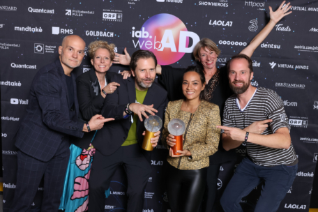 Sechs Personen posieren fröhlich vor einem Veranstaltungshintergrund, zwei von ihnen halten glänzende Preise in der Mitte. Andere gestikulieren aufgeregt in Richtung der Gewinner, die bei der iab.webAD-Veranstaltung alle lächeln und feiern.
