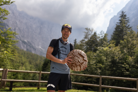 Ein Mann in sportlicher Kleidung steht im Freien und hält einen großen, runden, gemusterten Gegenstand in der Hand. Hinter ihm sind Holzzäune, grüne Bäume und hohe Berge unter einem teilweise bewölkten Himmel zu sehen.