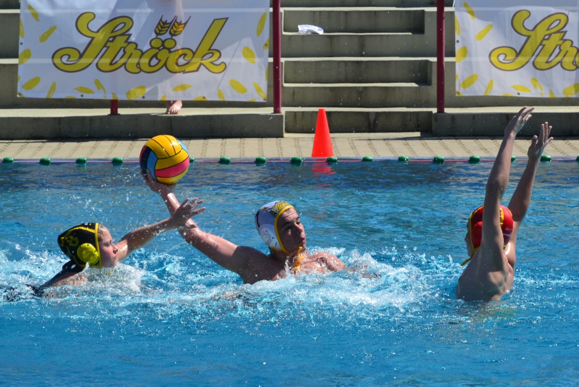 Drei Wasserballspieler treten in einem Schwimmbecken gegeneinander an; einer hält den Ball in der Mitte des Wurfs, während zwei Verteidiger zum Block greifen. Im Hintergrund erscheinen Stufen und ein Banner mit der Aufschrift "Ströck".