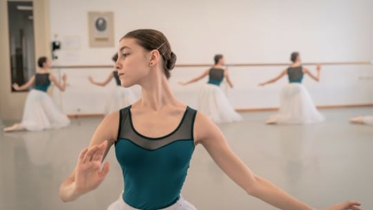 Eine junge Ballerina in einem blaugrünen Trikot und einem weißen Tutu posiert anmutig im Vordergrund, während andere Tänzerinnen in einem hellen, geräumigen Ballettstudio an der Stange üben.