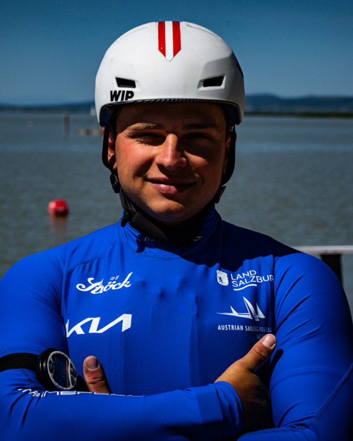 Ein Matrose in blauer Uniform und mit weißem Helm steht mit verschränkten Armen vor einem Gewässer.