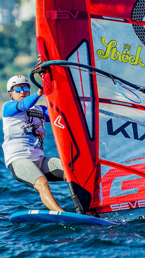 Ein Windsurfer, der einen Helm und eine blau getönte Schwimmbrille trägt, steuert ein großes rotes Segel über das Wasser.