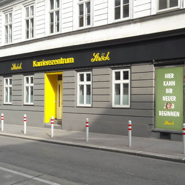 Ein graues Gebäude mit weißer Verkleidung hat eine leuchtend gelbe Tür und ein Schild mit der Aufschrift Karrierezentrum Ströck. Auf einem grünen Plakat im Fenster steht: "Hier kann Ihr neuer Anfang".