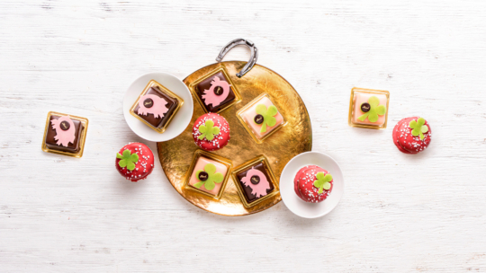 Auf einem goldenen Tablett liegen dekorative quadratische Kekse mit rosa und braunem Zuckerguss, daneben kleine runde rote Gebäckstücke mit grüner Verzierung; einige Leckereien sind auf weißen Schalen und einer weißen Holzfläche platziert.