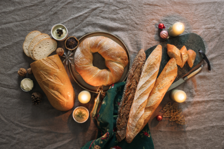 Verschiedene Brotsorten, darunter Baguette, ein runder Laib und geschnittenes Brot, werden auf einem Tisch mit Kerzen, Nüssen, einem grünen Tuch und festlichen Ornamenten arrangiert und schaffen eine gemütliche und einladende Atmosphäre.