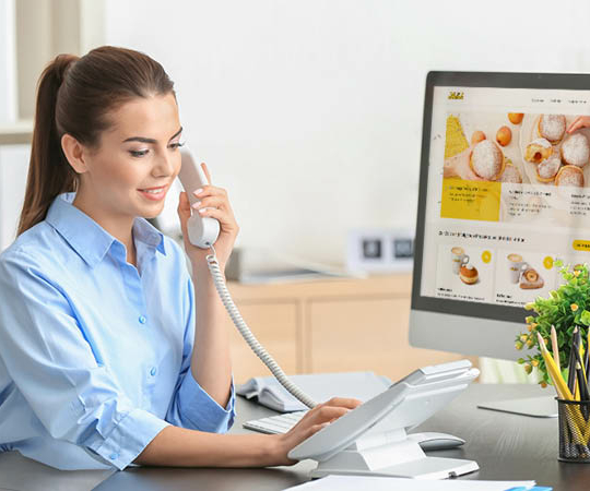 Eine Frau sitzt an einem Schreibtisch, telefoniert über ein Festnetztelefon und schaut auf ihren Computerbildschirm, auf dem die Website einer Bäckerei angezeigt wird. Auf dem Schreibtisch stehen eine Topfpflanze und Büromaterial.